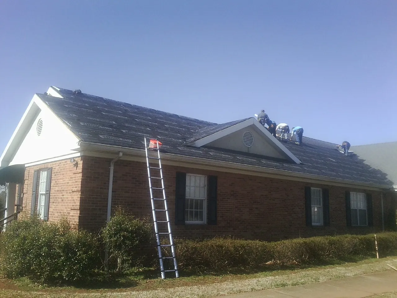 Crew tearing shingles off a brick ranch roof with a ladder set — Emergency Roof Repair work by our crew in Ellenton