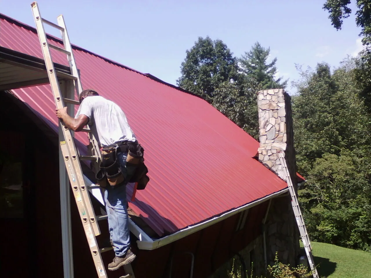 Roofer on a ladder beside a red metal roof and stone chimney — Soffit & Fascia Repair by Roofing Brothers in Ellenton